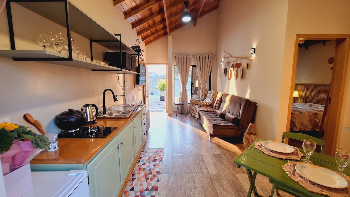 A kitchen area is seen, featuring light green cabinetry and a wooden countertop. A seating area includes a sofa and a small dining table set for two. The space is illuminated by natural light from large windows, and decorative elements adorn the walls.