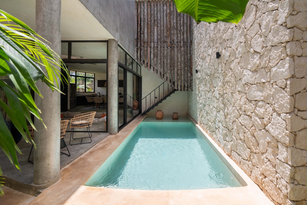 A private pool is surrounded by natural stone walls and lush greenery, creating a serene atmosphere. The space features sleek, modern lines with large glass doors leading to the living area. Terracotta pots decorate the area, enhancing the connection to the outdoors.