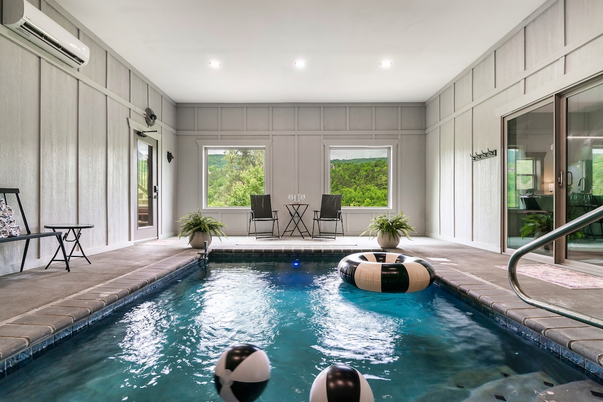 An indoor heated pool is illuminated with bright overhead lights. Two lounge chairs are positioned near the shallow end, while large windows provide views of the greenery outside. A comfortable swimming area features floating pool toys, and steps lead to the entry.