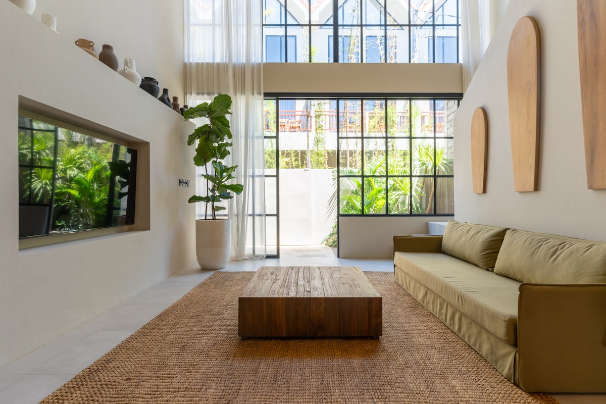 A spacious living area features a large, light-filled window wall, offering views of lush greenery. A low wooden coffee table sits on a natural fiber rug, complemented by a comfortable sofa in muted tones. Minimalist decor is accentuated by wooden wall art.