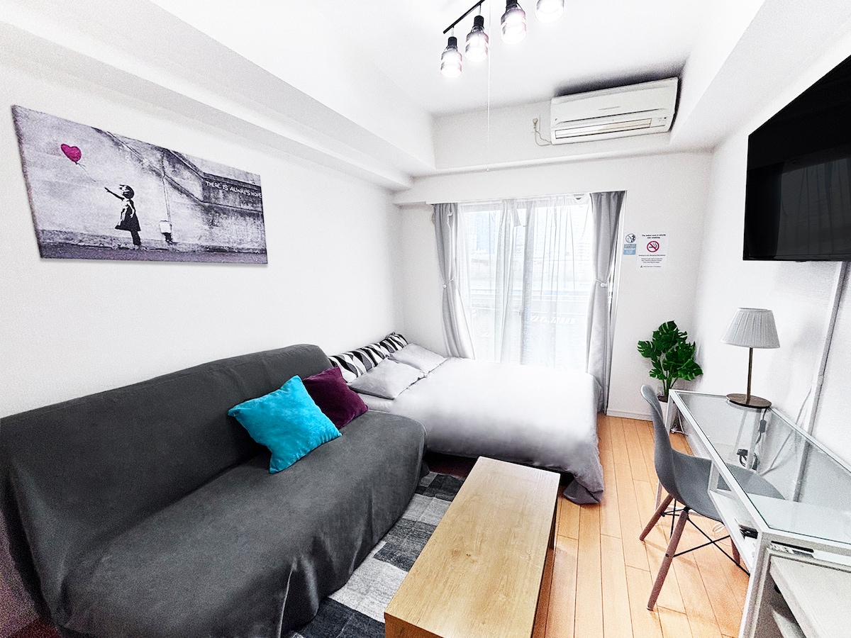 The living area exhibits a modern design with a gray sofa adorned with colorful cushions. A spacious bed with soft bedding is positioned beside the large window, partially draped with light curtains. A small wooden coffee table sits in the center, accompanied by a clear desk and plant.