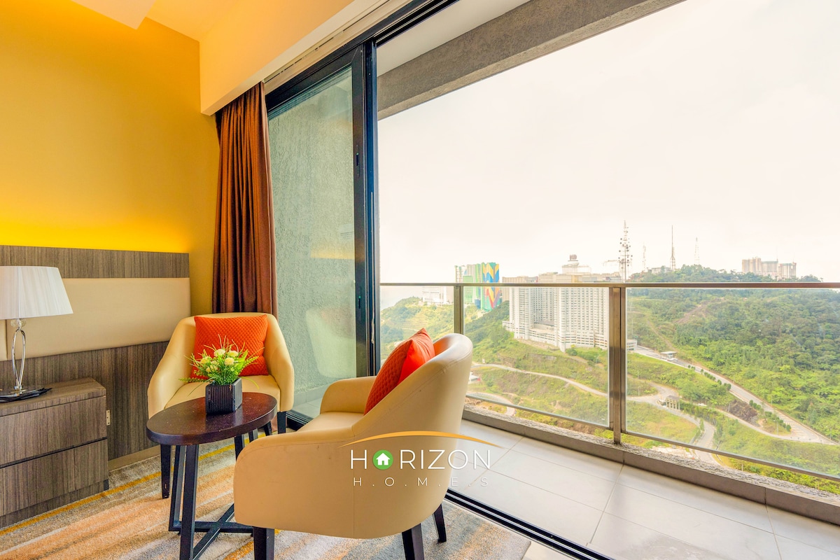 A seating area is showcased, featuring two orange accent chairs positioned near a large glass window. A small table is placed beside the chairs, complementing a modern lamp on an adjacent nightstand. The view outside reveals green hills and urban structures.