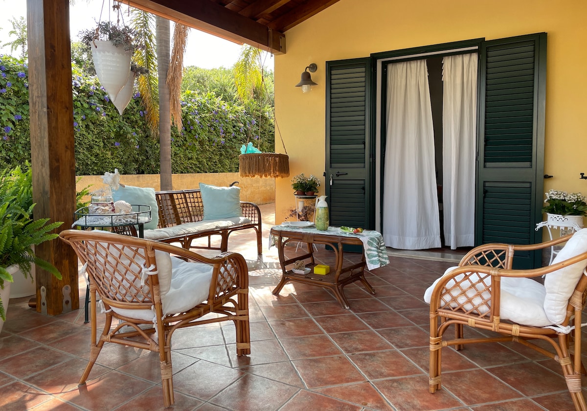 A covered outdoor seating area is featured, furnished with multiple rattan chairs and a coffee table. Lush greenery serves as a backdrop, creating a serene environment. Soft cushions adorn the chairs, while natural light filters through the open shutters, enhancing the inviting space.