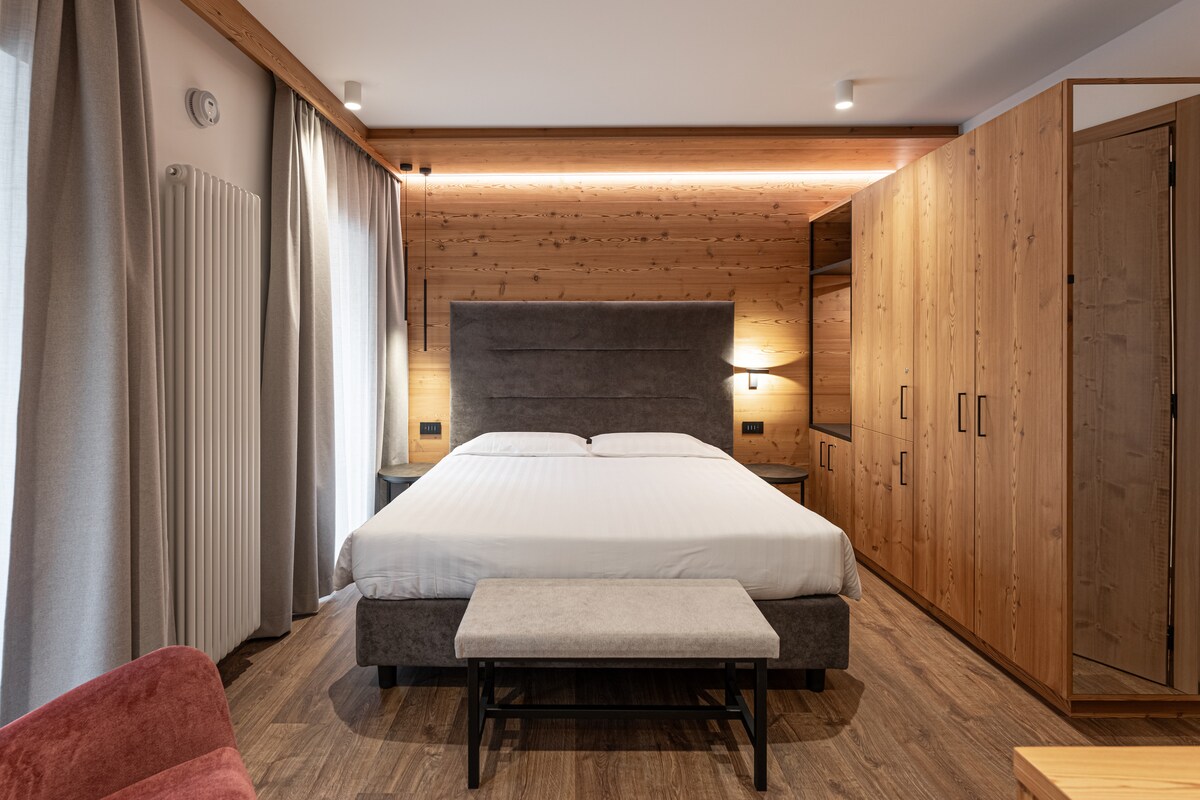 A spacious room with wooden wall paneling, featuring a comfortable bed centered between two windows. A seating area includes a small bench at the foot of the bed. A wardrobe and mirror provide additional storage and functionality, along with soft lighting along the top edge.