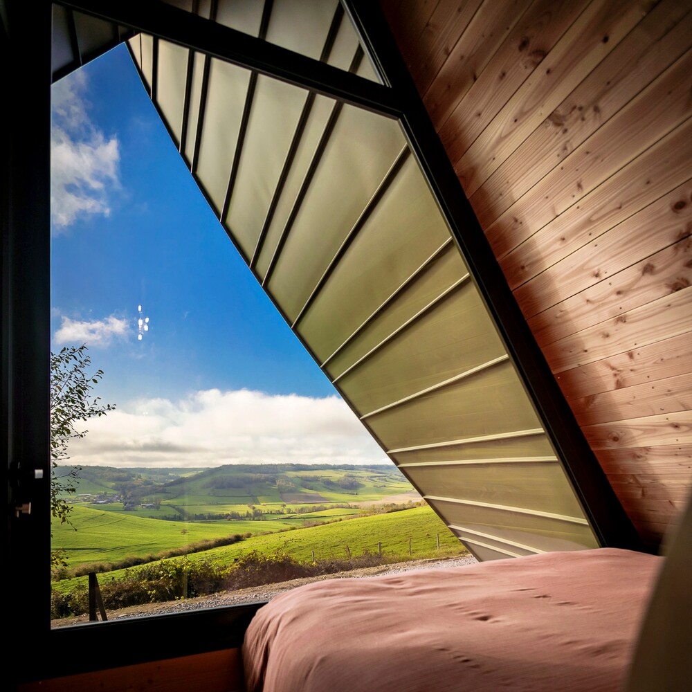A large window features a unique triangular design, offering views of expansive green hills and distant valleys. Soft natural light fills the room, highlighting smooth wooden walls and a spacious bed, creating a serene atmosphere for guests.