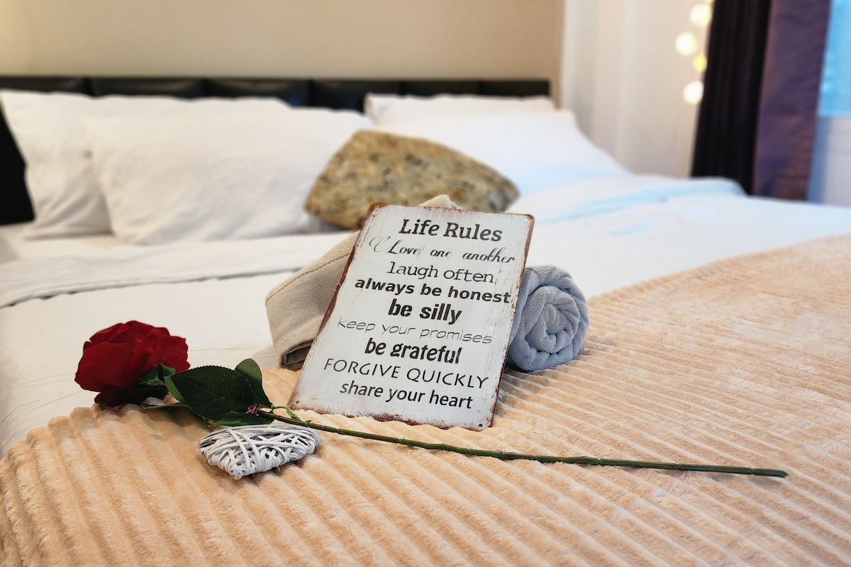 A cozy bed is adorned with a light-colored blanket, accentuated by decorative items including a framed sign with inspiring phrases. Two roses and a rolled towel are placed elegantly on the bedding, adding thoughtful touches to the inviting space.