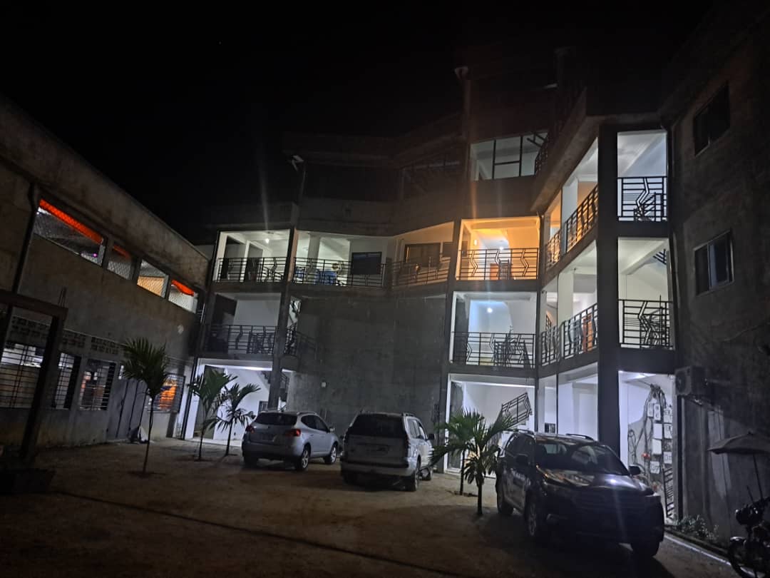 The image showcases a modern apartment building illuminated at night, featuring multiple floors with large windows. Vehicles are parked in the courtyard, surrounded by palm plants. The structure's contemporary design emphasizes open spaces and balancing light and shadow.