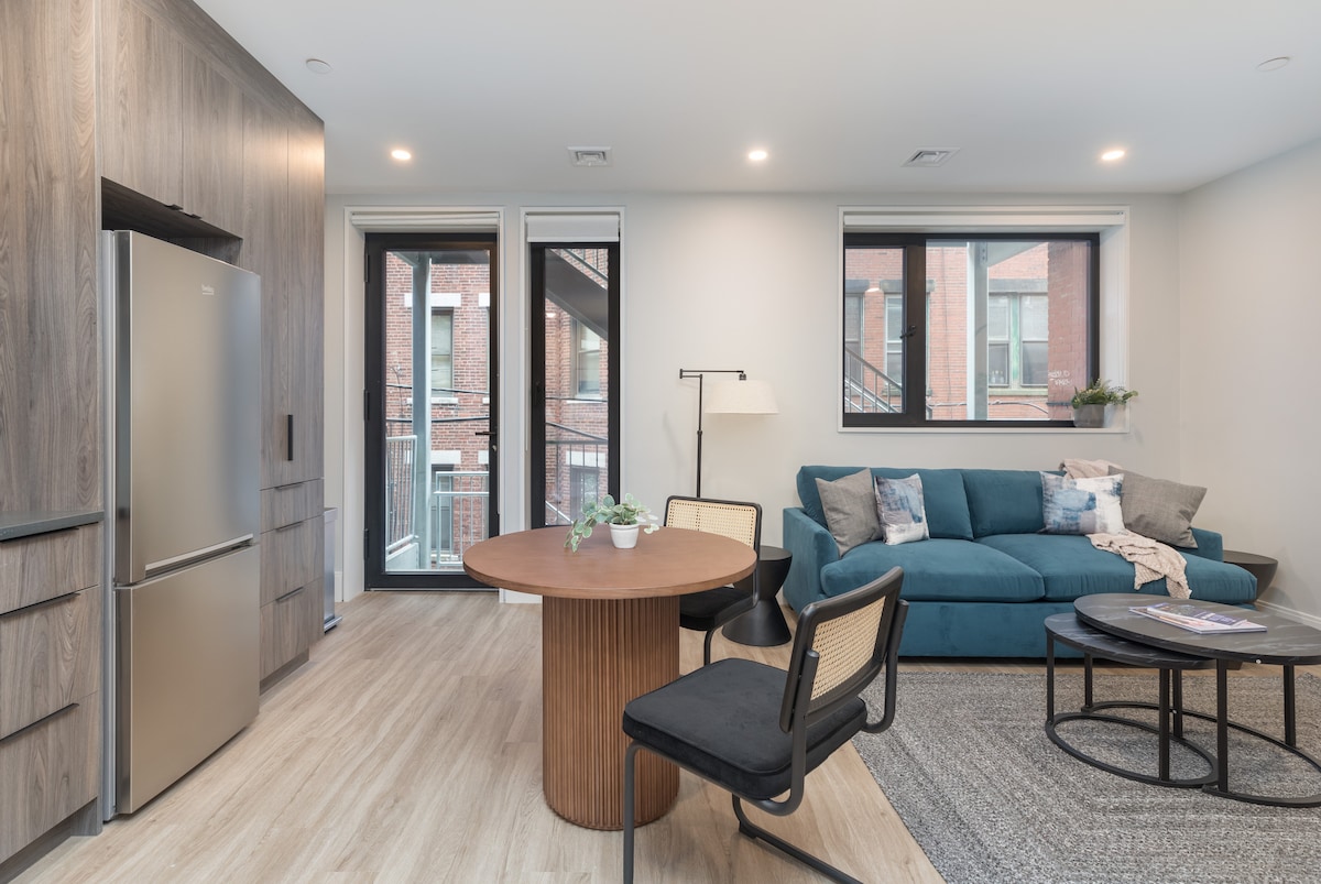 A modern living area showcases a stylish blue couch paired with two black chairs and a round wooden dining table. Large windows provide natural light, revealing views of the outdoor staircase. A sleek stainless steel refrigerator is positioned against the wall, enhancing the contemporary design.