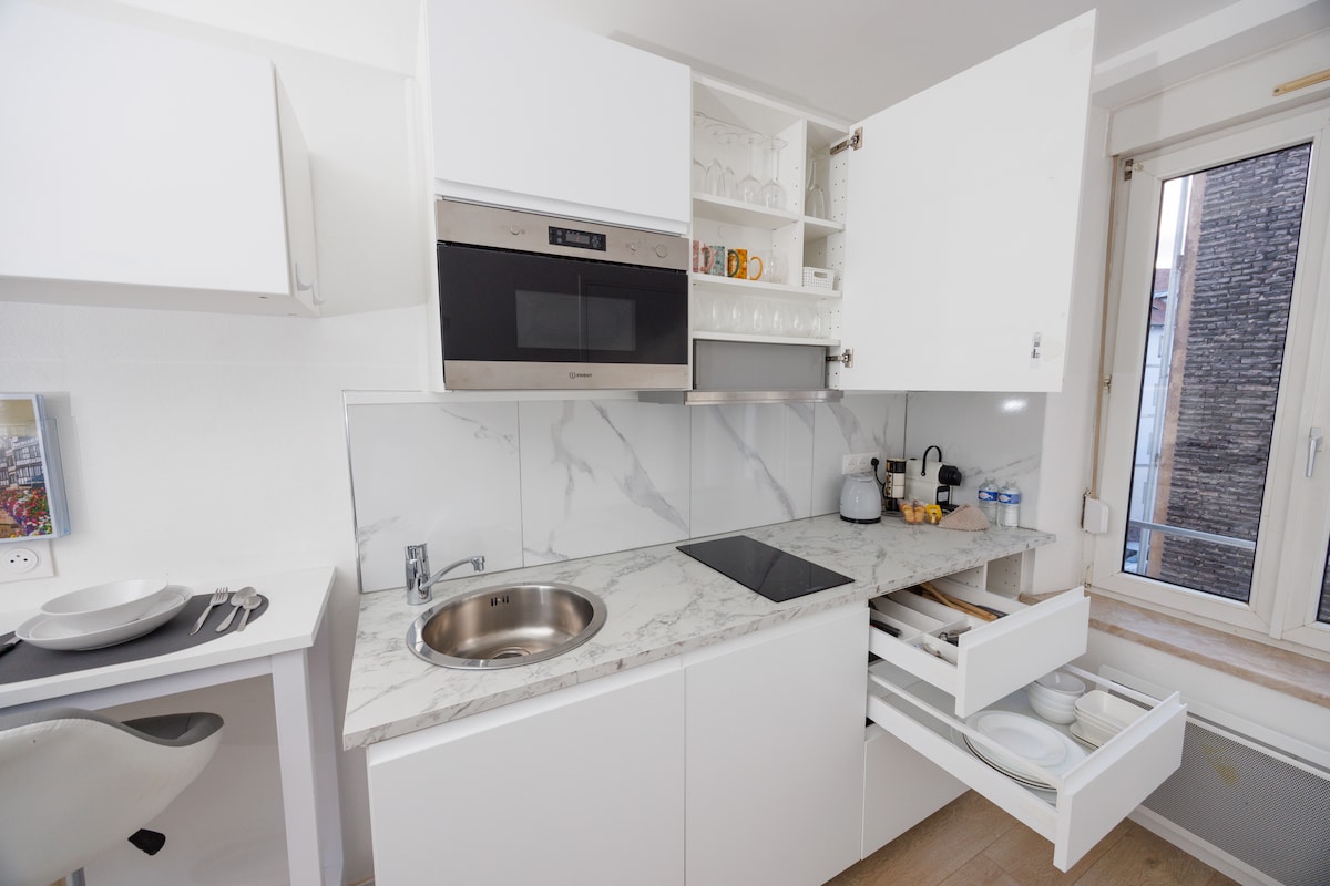 A modern kitchen space is displayed with sleek white cabinetry and marble countertops. An integrated oven and microwave are visible, alongside a hot plate. Drawers are partially open, showcasing dishware, while a window allows natural light to illuminate the area.