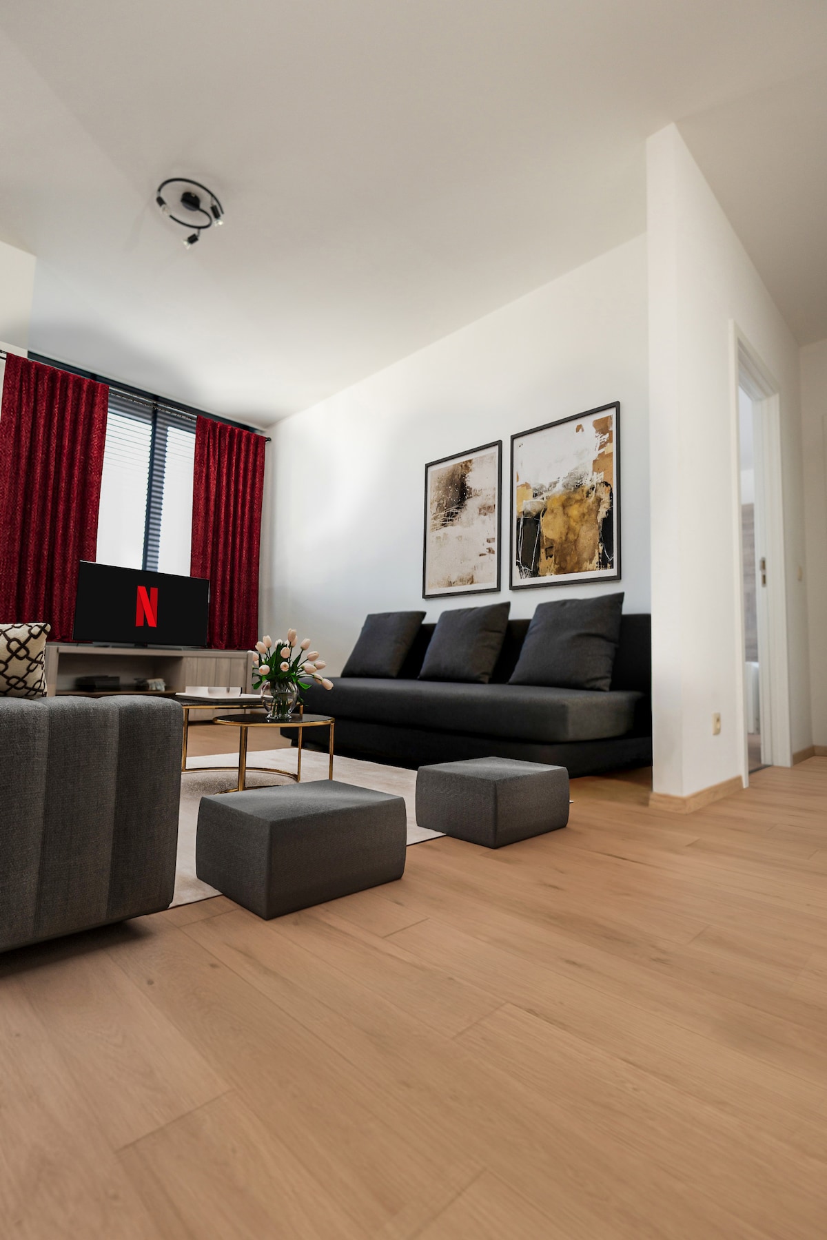 A modern living room features a dark gray sofa and two matching ottomans, positioned beside a low coffee table. Two artistic prints adorn the light-colored walls, while vibrant red curtains frame a window, allowing natural light to brighten the space.