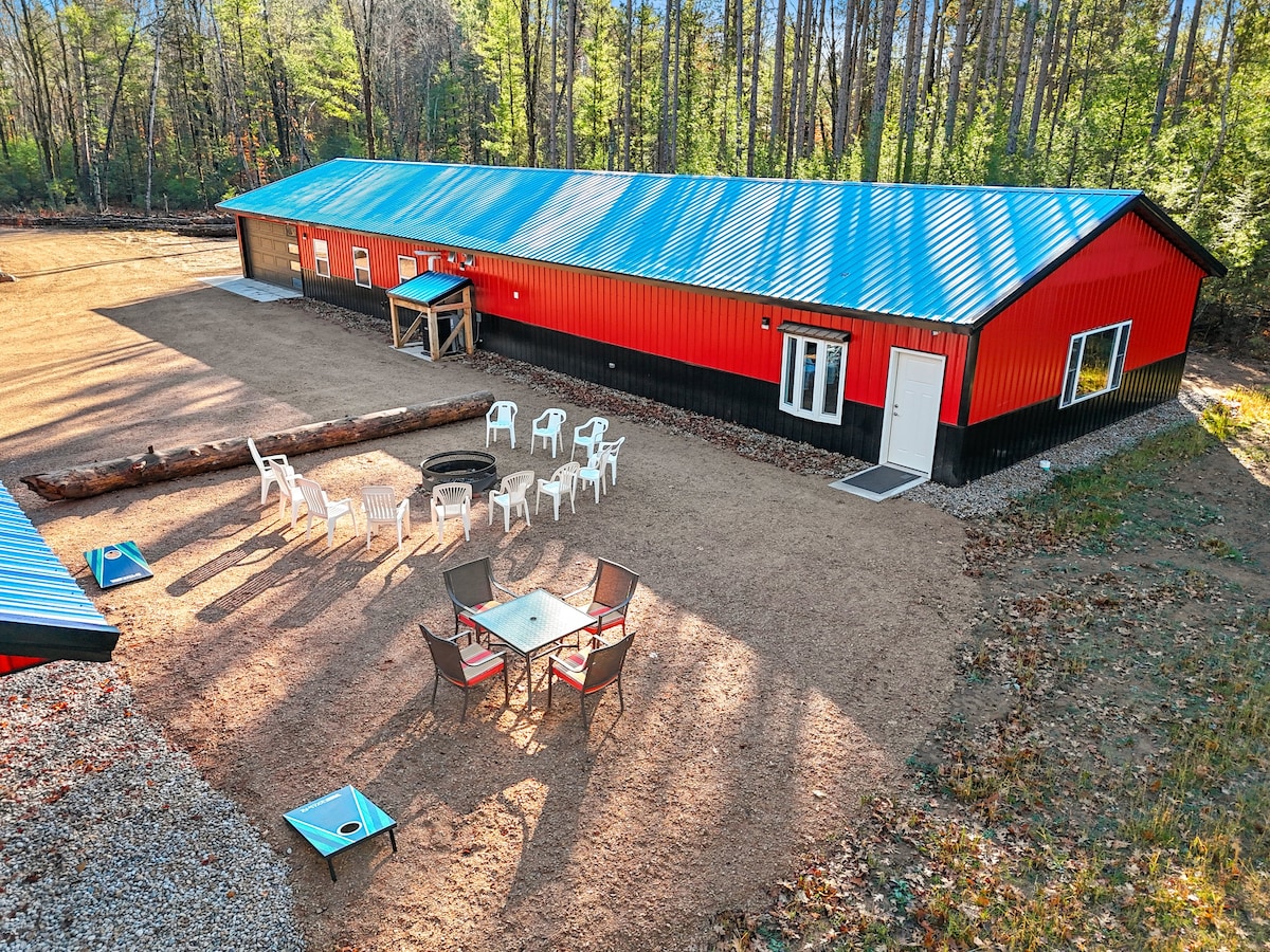 The exterior of a vibrant red building is paired with a blue metal roof, surrounded by trees. A gravel area features a circular arrangement of white chairs, a fire pit at the center, and a shaded seating space with a table and chairs, creating an inviting outdoor gathering spot.