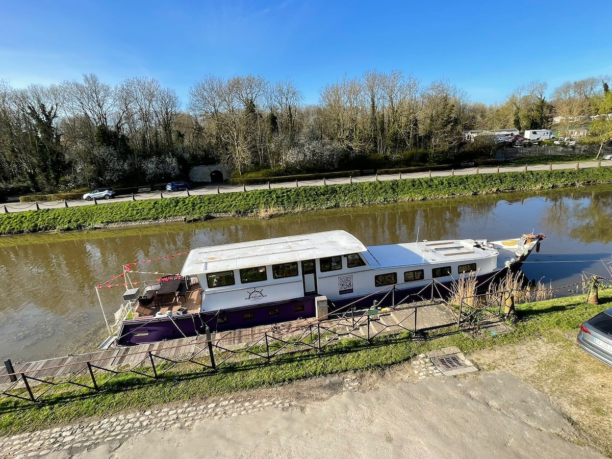 A moored houseboat is positioned alongside a tranquil canal, surrounded by greenery. The boat features an outdoor seating area and is accessible via a wooden walkway. Bright blue skies contrast with the calm water, enhancing the peaceful ambiance of the location.