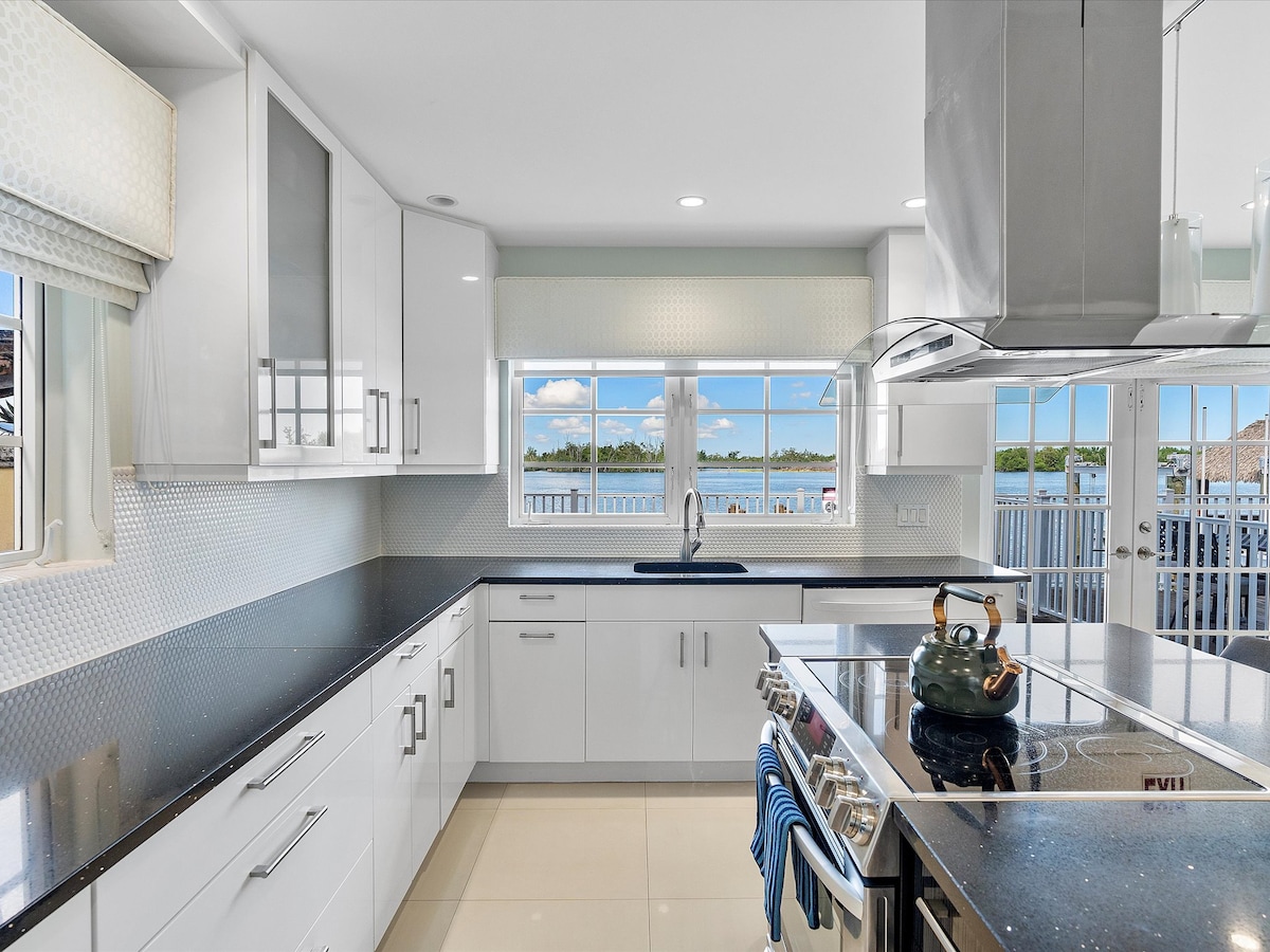A modern kitchen is presented with sleek white cabinets and dark countertops. A stainless steel stove and hood are central to the space, while large windows allow natural light to illuminate the area. Scenic views of the water are visible through the glass panes.
