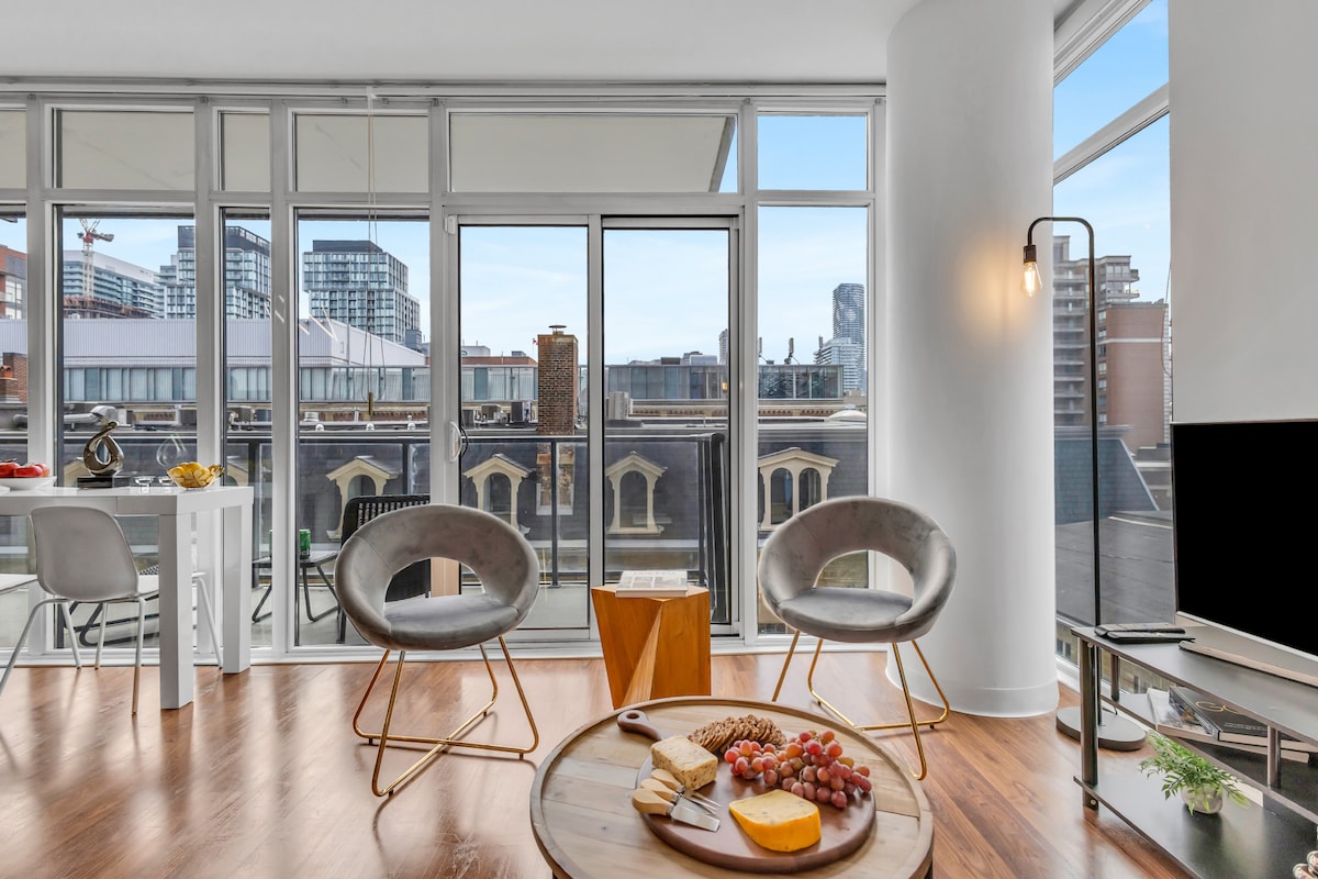 A bright living space features large glass windows offering views of the city skyline. Two sleek chairs are positioned around a circular coffee table with a cheese and fruit platter. A dining table for two is visible in the background, enhancing the room's charm.