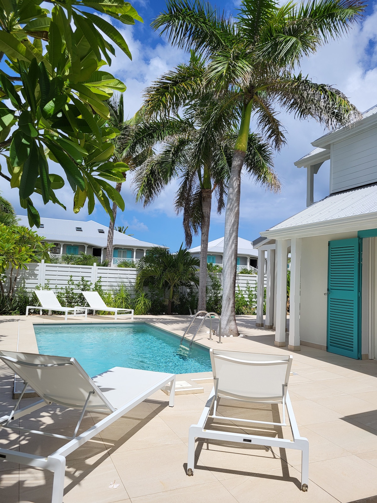 A refreshing pool area is surrounded by tropical palm trees, providing a serene outdoor setting. Two white lounge chairs are positioned by the pool, inviting relaxation under the clear blue sky. Bright turquoise doors of the villa can be seen in the background, enhancing the inviting atmosphere.