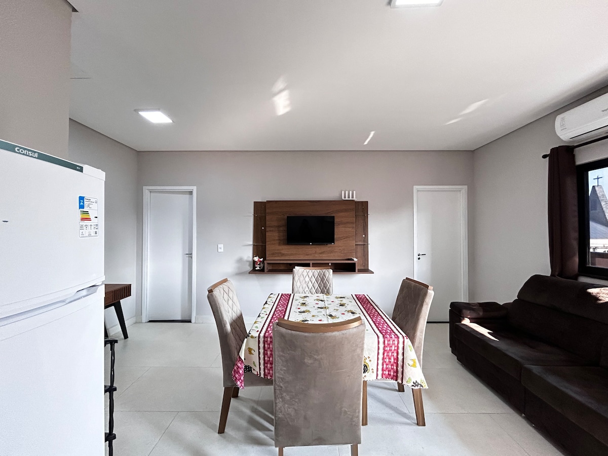 The living and dining area features a large table surrounded by four chairs, complemented by a dark brown sofa. A mounted television is positioned on the wall opposite the sofa, while natural light enters through the windows, illuminating the neutral-colored walls.