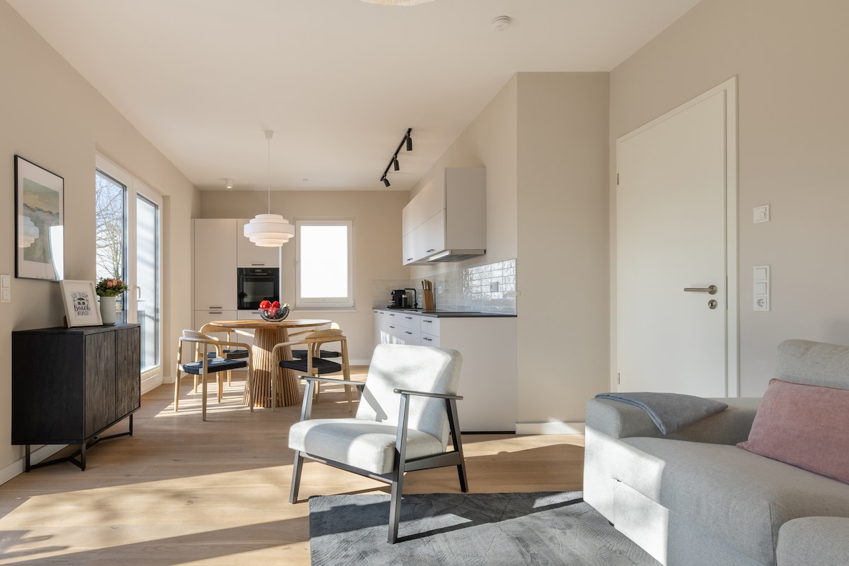 A modern living space features a light-filled kitchen and dining area with a round wooden table and four chairs. Neutral-colored walls and furnishings enhance the openness. Light wood flooring and a small black cabinet add warmth, while large windows invite natural light.