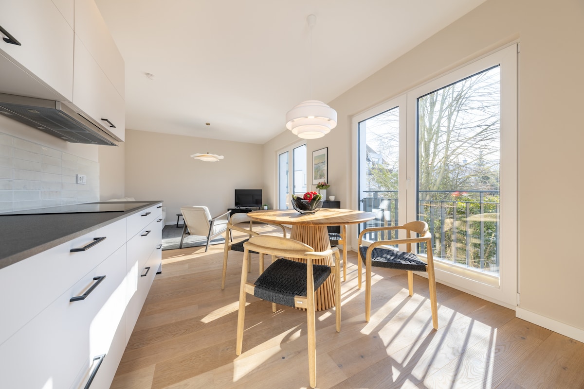 A bright dining area features a round wooden table surrounded by four chairs with woven seats. Large windows invite natural light, revealing a view of greenery outside. The simple kitchen cabinetry provides a clean and modern look, complemented by a cozy seating arrangement in the corner.