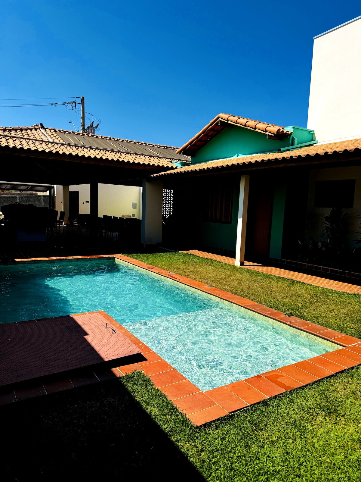 A clear swimming pool is surrounded by manicured grass, with a tiled deck leading to an overhanging roof that provides shade. Bright blue skies and a well-maintained exterior of the villa contribute to a relaxing outdoor environment.