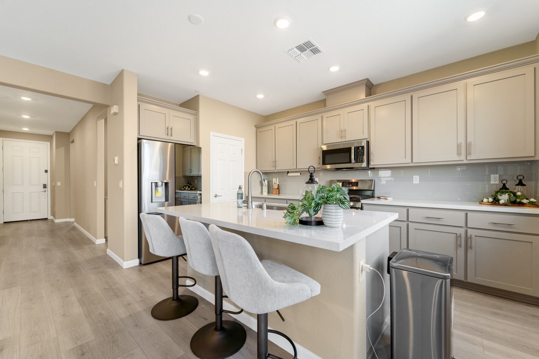A modern kitchen features sleek cabinetry and granite countertops. Bar seating with plush stools is available at the island, while stainless steel appliances complement the neutral color palette. Natural light fills the space through a nearby entrance, enhancing the inviting atmosphere.