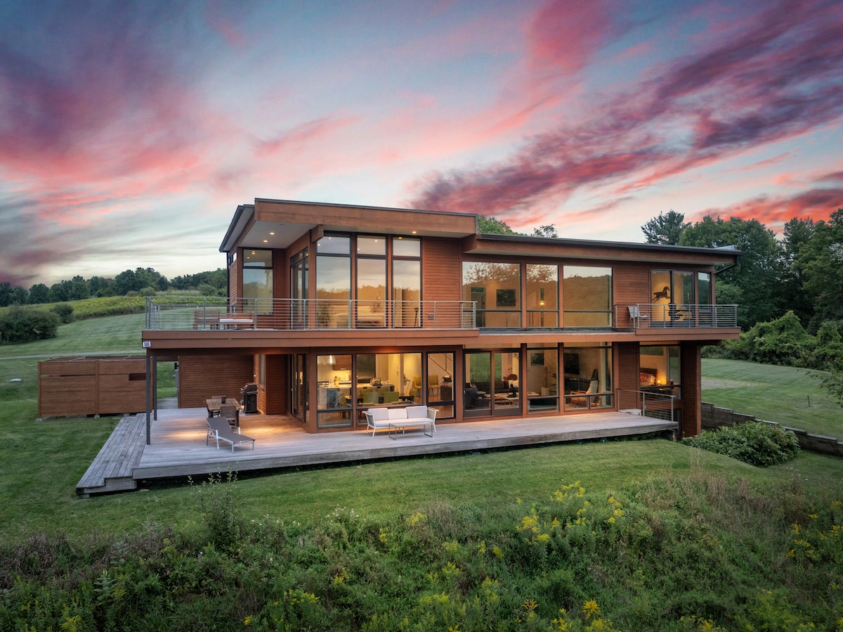 The modern lakefront home features a sleek exterior with large windows reflecting the stunning sunset. A spacious deck extends from the house, furnished with seating that overlooks a lush green landscape, creating a relaxing outdoor environment.