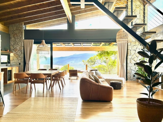 An open living space with large glass doors allows natural light to flood the area. A spacious leather sofa faces a wooden dining table, surrounded by chairs. The kitchen area features stone accents. A staircase is visible in the background, leading to an upper level.