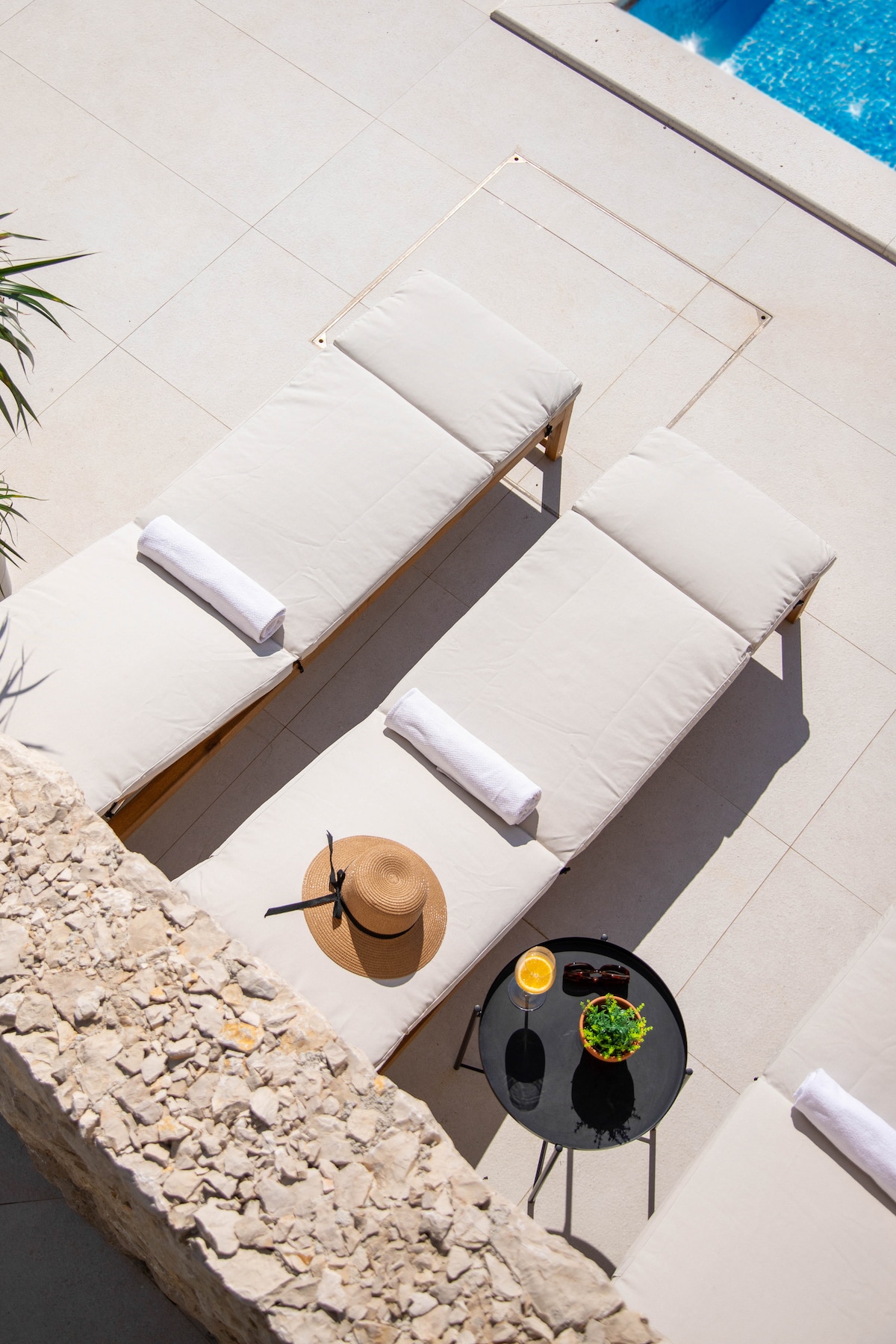 Two sun loungers are arranged beside an outdoor swimming pool, featuring light-colored cushions and neatly placed towels. A small black table holds a refreshing beverage and a potted plant. A wide-brimmed hat rests on one of the loungers, emphasizing a relaxed outdoor setting.