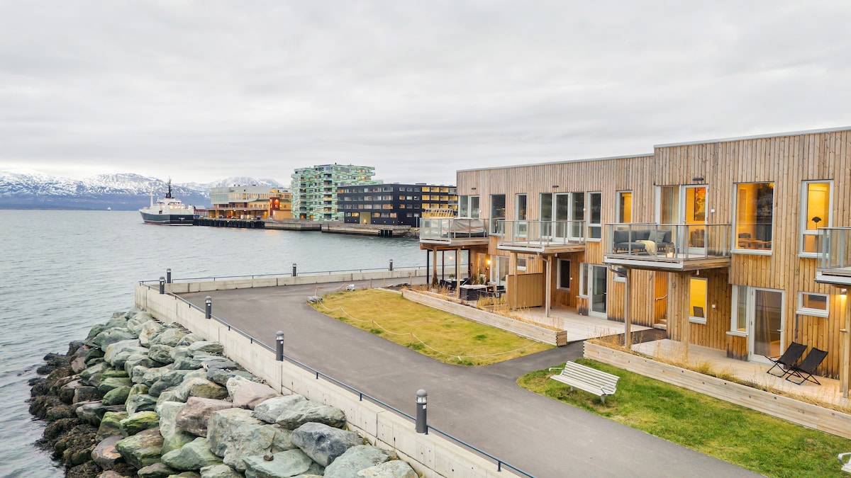 A modern two-story townhouse is positioned along a waterfront, featuring large windows and balconies that offer views of the sea. A grassy area is bordered by a pathway, while large stones line the shore. Nearby buildings reflect a contemporary architectural style.