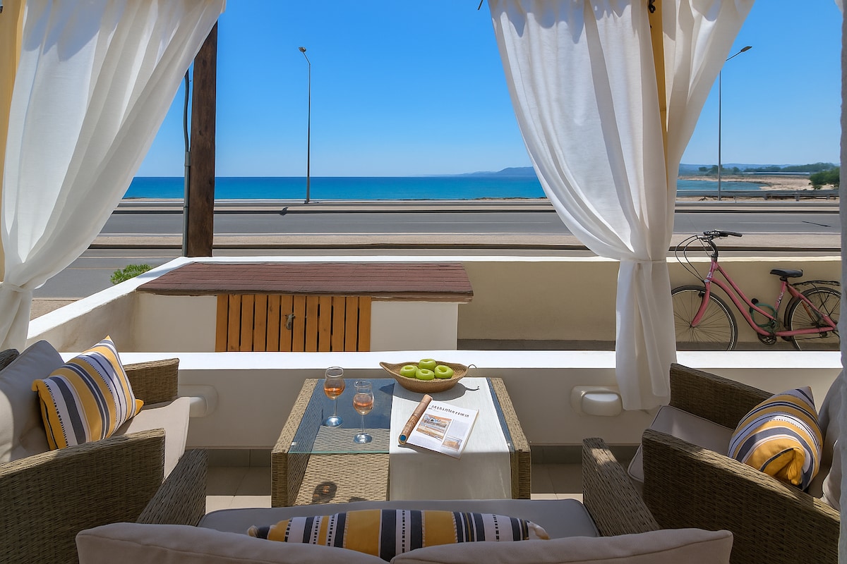 A front patio is depicted with two stylish chairs and a glass-top coffee table. A small plate with sliced kiwi and two glasses sits on the table. Beyond, the inviting view of the sea is visible, complemented by a clear blue sky.