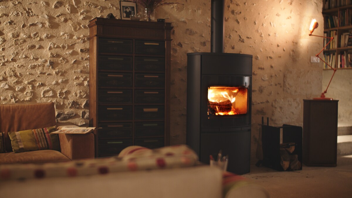 A wood-burning stove is situated against a textured stone wall, providing warmth to the cozy interior. A vintage style cabinet is adjacent, accompanied by a plush sofa adorned with multi-colored cushions. Soft lighting enhances the tranquil atmosphere of the living space.