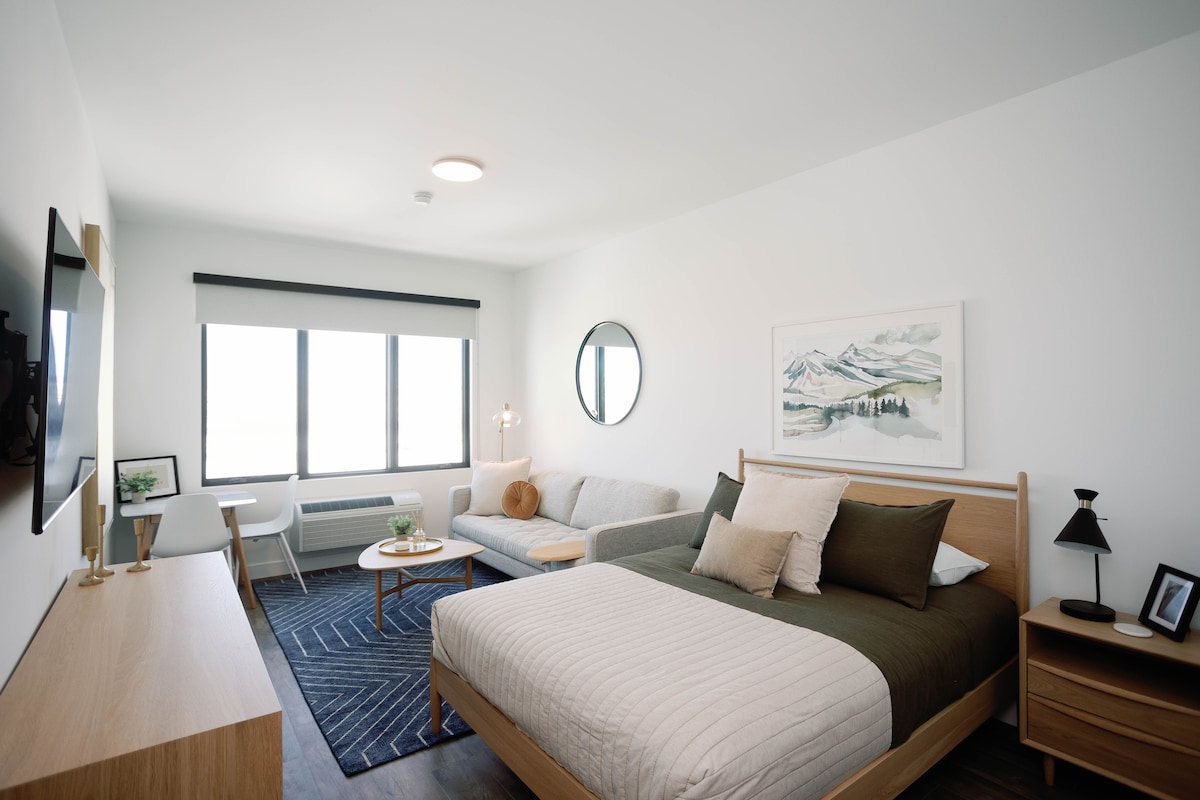 A studio room features a spacious bed with earthy-toned bedding, complemented by a cozy couch and a round coffee table. Large windows allow natural light to fill the space, while a decorative wall art piece and modern furnishings create a relaxed environment.
