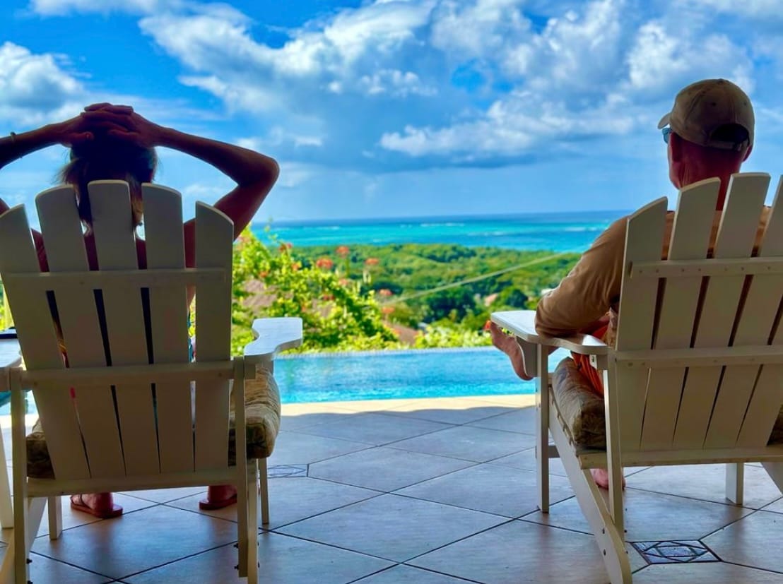 Two comfortable chairs are positioned on a tile patio, overlooking a scenic ocean view. Lush greenery surrounds the area, and a vibrant blue sky with scattered clouds enhances the relaxing atmosphere. The infinity pool is visible, seamlessly blending with the horizon.