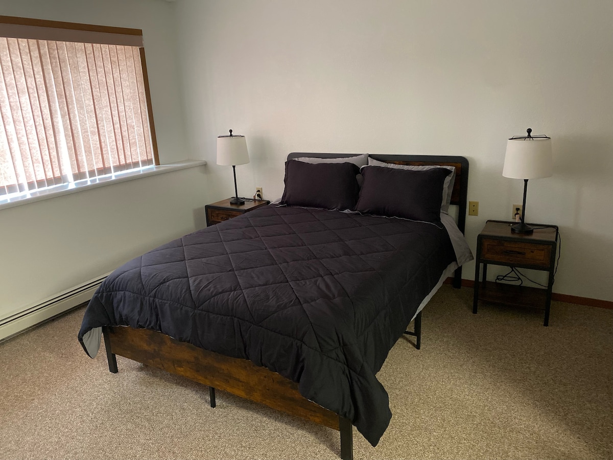 A spacious bedroom features a queen bed dressed in dark bedding. Two bedside tables with lamps are positioned on either side. Natural light filters through the window, where vertical blinds provide privacy.