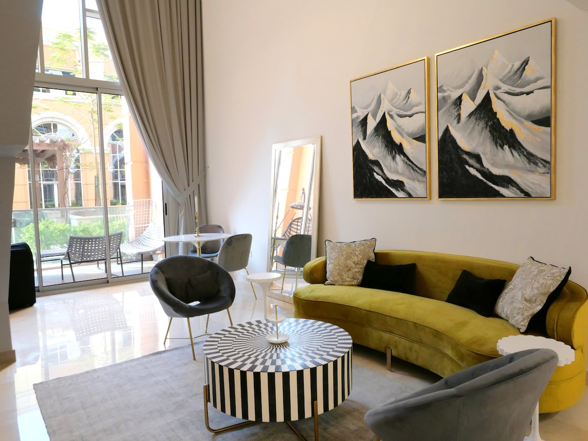 A spacious living area features a yellow curved sofa complemented by gray armchairs and a striped coffee table. Two large mountain-themed artworks adorn the wall, while natural light flows through the expansive glass doors, showcasing views of the outdoor space.