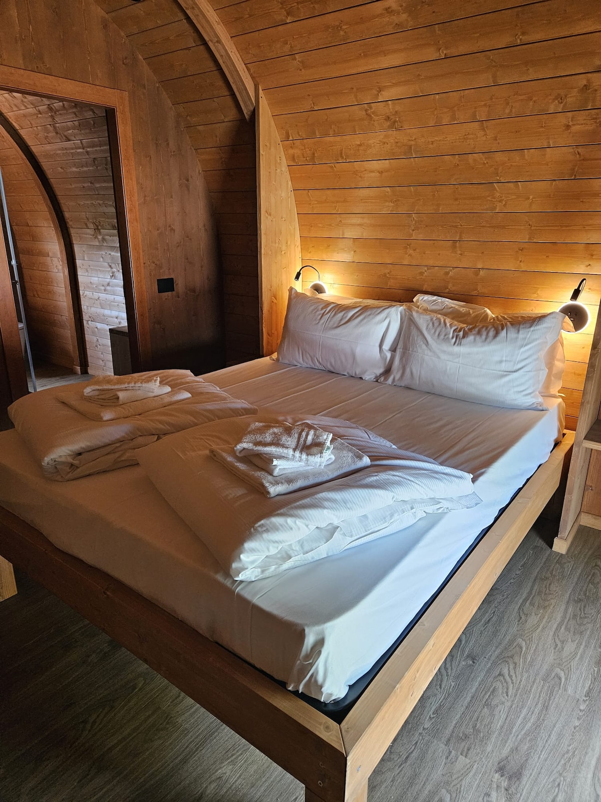 A cozy sleeping area is highlighted by wooden walls and an arched ceiling. A spacious bed is adorned with neatly arranged pillows and soft linens, creating a serene environment. Warm lighting from bedside fixtures adds a touch of comfort to the atmosphere.