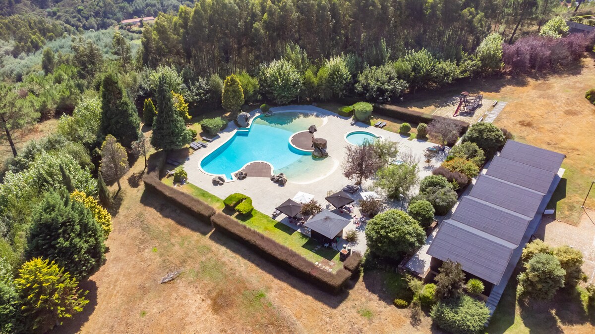 The aerial view reveals a spacious outdoor area featuring a large, inviting swimming pool surrounded by landscaped gardens. Sun loungers and shaded seating areas are visible, along with varied greenery and trees providing a natural setting. The layout suggests ample space for relaxation and leisure activities.