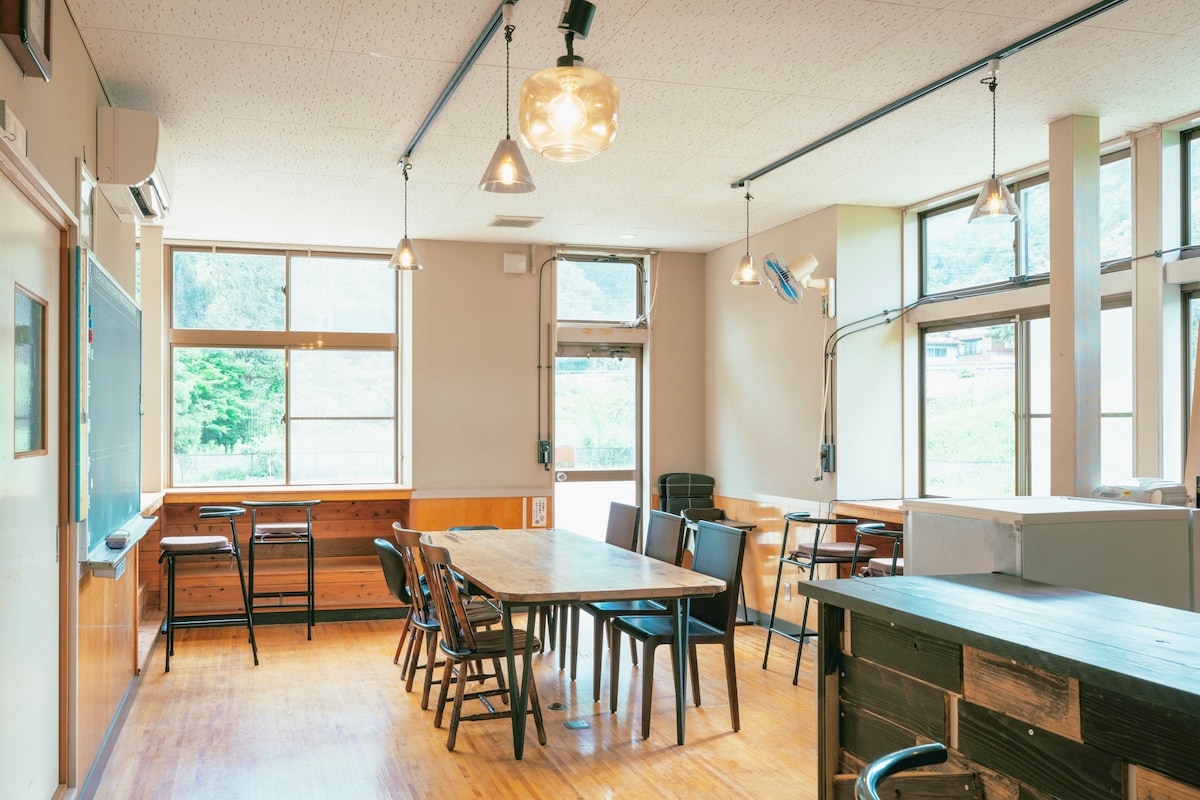 An open common area features large windows that provide natural light, illuminating the wooden flooring. A spacious wooden table is surrounded by a mix of chairs, and additional seating is arranged near the walls. Subtle lighting fixtures hang from the ceiling, enhancing the room's functionality.