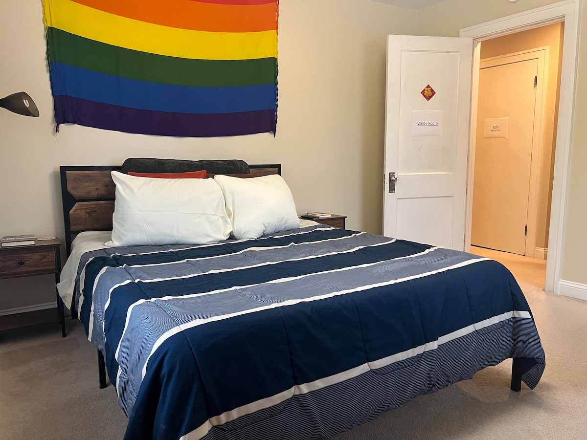 A spacious private room is shown, featuring a queen bed with a navy and white striped comforter. Adjacent to the bed, a bedside table holds a lamp. A rainbow flag is displayed on the wall above the bed, and a door leads to the adjacent bathroom.