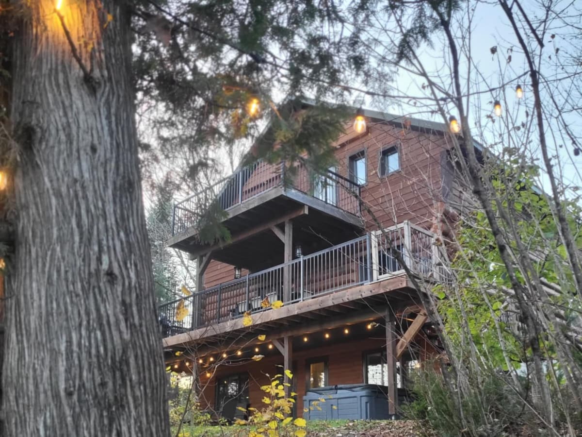 A rustic log cabin is set against a backdrop of trees, showcasing a spacious porch with railing. The hot tub is visible on the lower deck, surrounded by natural foliage. Warm lights are strung along the structure, enhancing the cabin's inviting exterior.