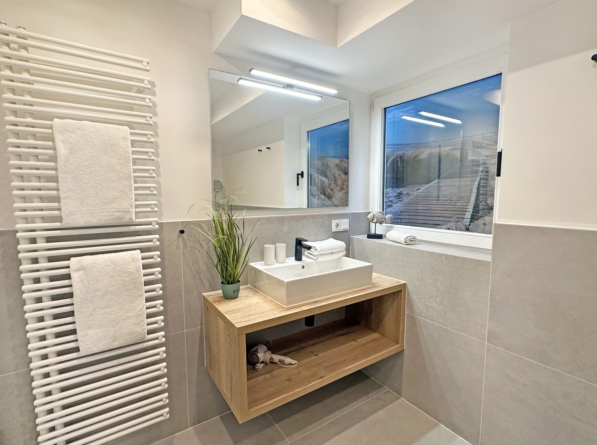 A modern bathroom features a wooden shelf with a sink and toiletries. Two bath towels are neatly hung on a heated towel rack beside a large mirror. Natural light enters through two windows, illuminating the space with a fresh and clean ambiance.