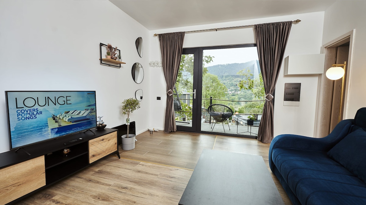 A modern lounge area features a plush blue sofa paired with a black coffee table. A large television is positioned against a light wood entertainment unit. Expansive windows allow sunlight to fill the space, providing a view of the natural surroundings and access to a small outdoor balcony.