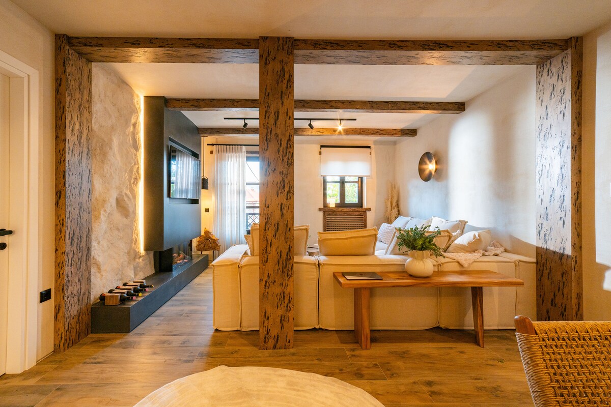 A cozy living area features a sectional sofa with soft cushions, complemented by natural wooden beams that frame the space. A decorative fireplace is positioned against a dark wall, while a wooden coffee table rests in front, creating a harmonious and inviting ambiance.