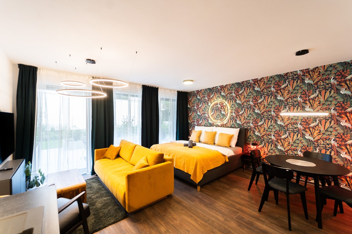 The room features a spacious sleeping area highlighted by a comfortable bed with soft linens. A vibrant yellow sofa adds a touch of freshness, alongside a modern dining table with chairs. Large windows allow natural light to illuminate the floral wallpaper, enhancing the overall ambiance.
