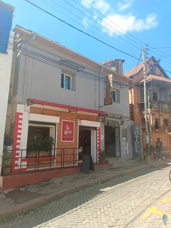 A two-story building is featured, with a decorative facade in light grey and warm beige tones. A small outdoor seating area is visible, along with a sign displaying the name of the establishment. The surrounding street is lined with other colorful buildings.