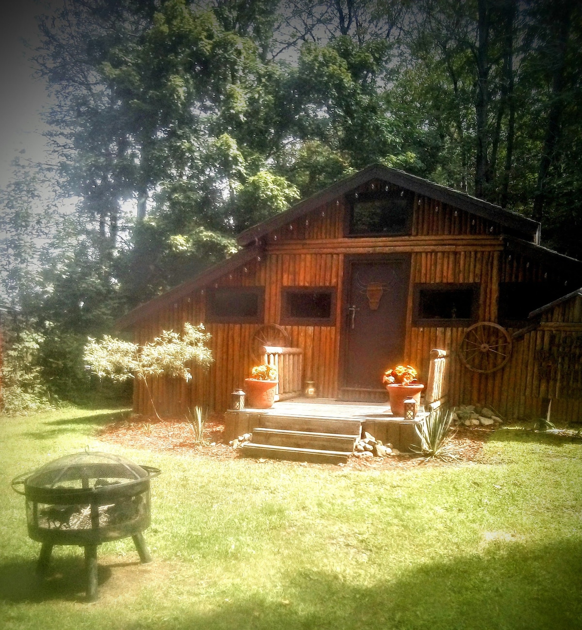 A rustic cabin showcases a wooden façade with large windows and a distinct roofline. The entrance is framed by flower pots, and steps lead to the door, which is accentuated by decorative elements. A fire pit sits in the foreground on a grassy area.