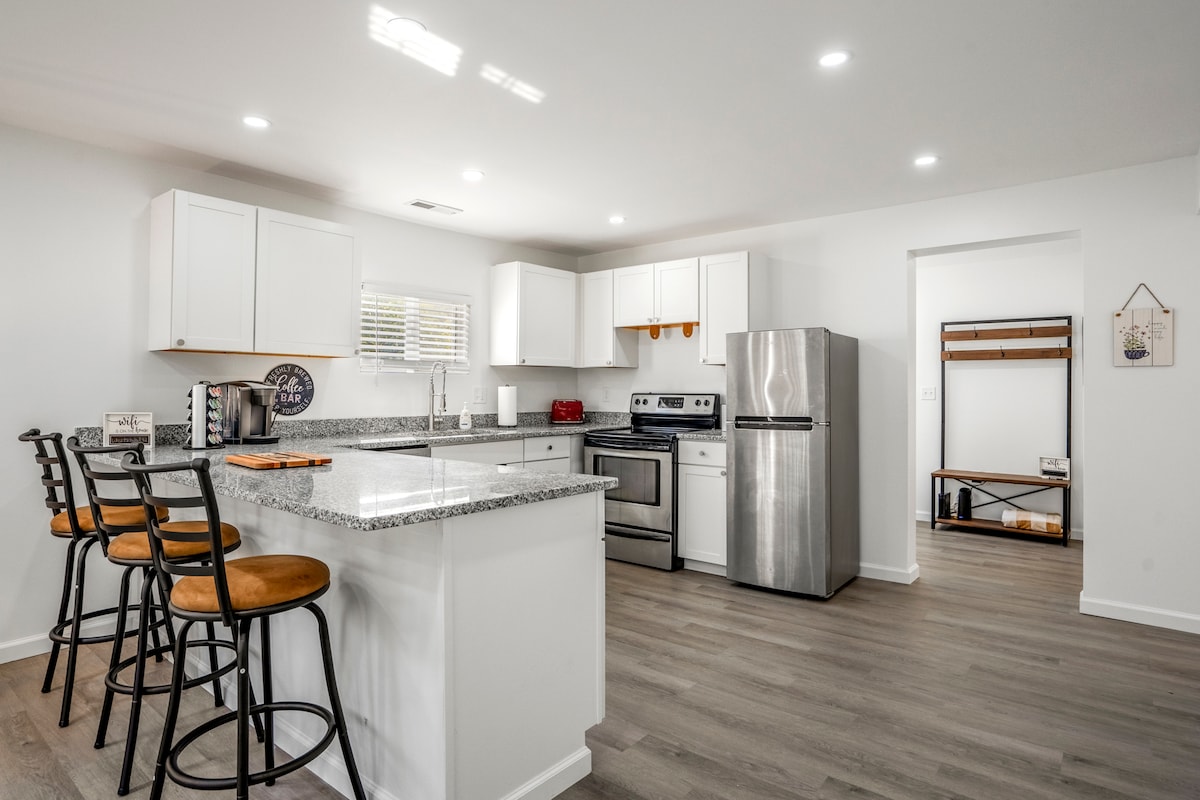 A modern kitchen features white cabinetry and stainless steel appliances. A spacious island with four bar stools is situated at one end, while natural light enters through a nearby window. The flooring adds a contemporary touch to the open layout.