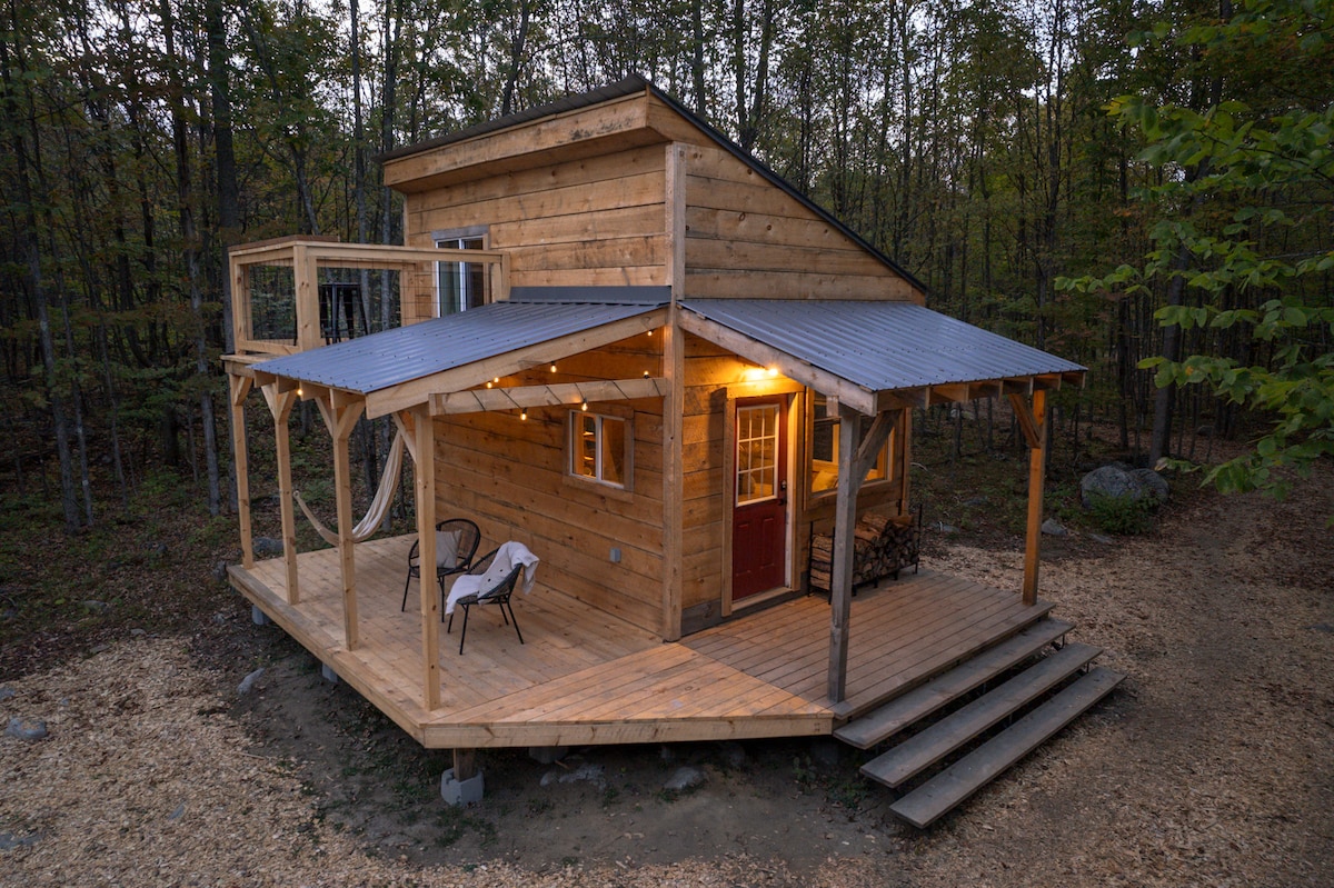 The exterior of the cabin showcases a wooden structure surrounded by trees, featuring a deck with a covered area. Two chairs are positioned on the deck, and a wood stack is visible nearby. The warm evening light enhances the welcoming ambiance of the cabin.