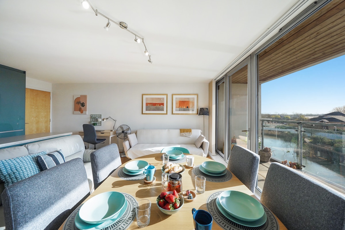 A spacious open-plan living area features a round dining table set with teal plates and glasses. The large glass doors offer views of the river and outdoor space. Soft seating is arranged nearby, creating an inviting atmosphere for meals and relaxation.