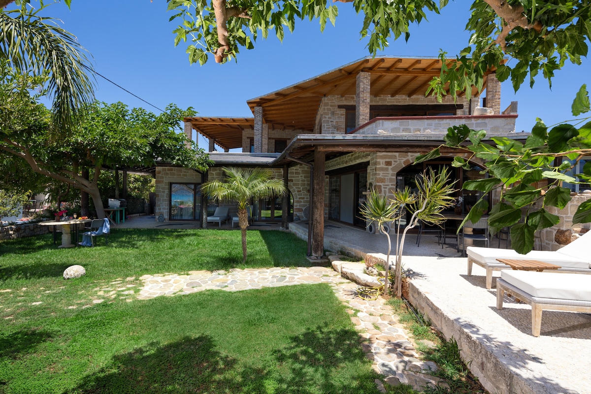 The exterior of the villa showcases a blend of traditional stonework and modern architecture. Lush greenery surrounds the outdoor space, featuring a well-maintained lawn and shaded seating areas. Sun loungers are positioned on the stone patio, inviting relaxation in the tranquil setting.