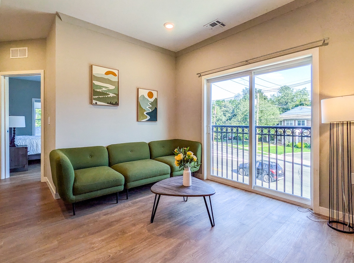 A cozy living area features a green sectional sofa paired with a round coffee table. Natural light filters through large sliding glass doors, which open to a balcony overlooking the street. Artwork hangs on the walls, adding a touch of personality to the space.